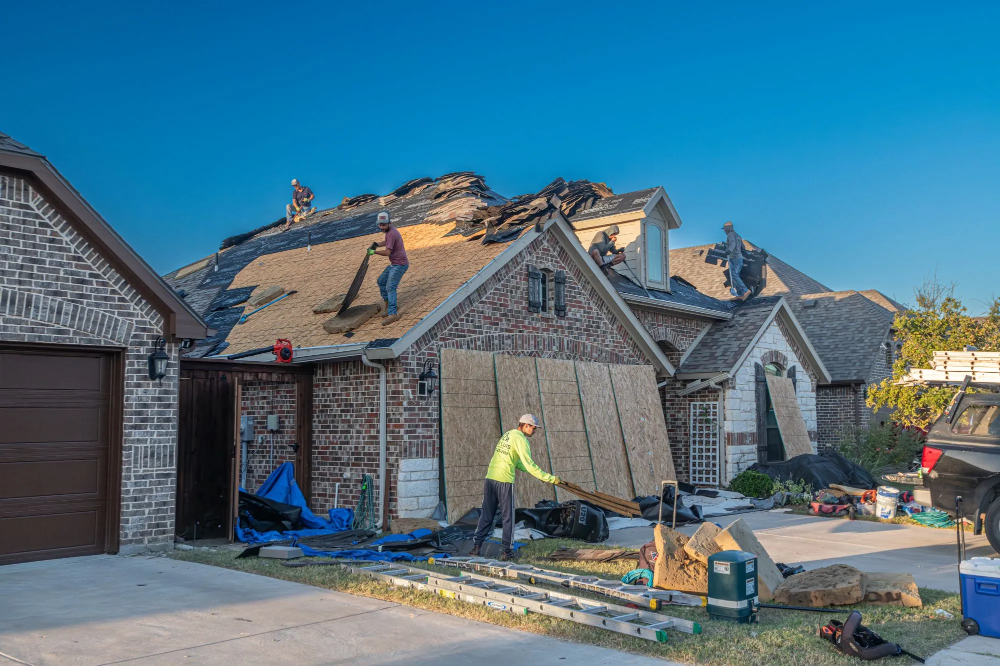 A roofing crew re-covering a large brick house, ladders up and the drive sheeted over