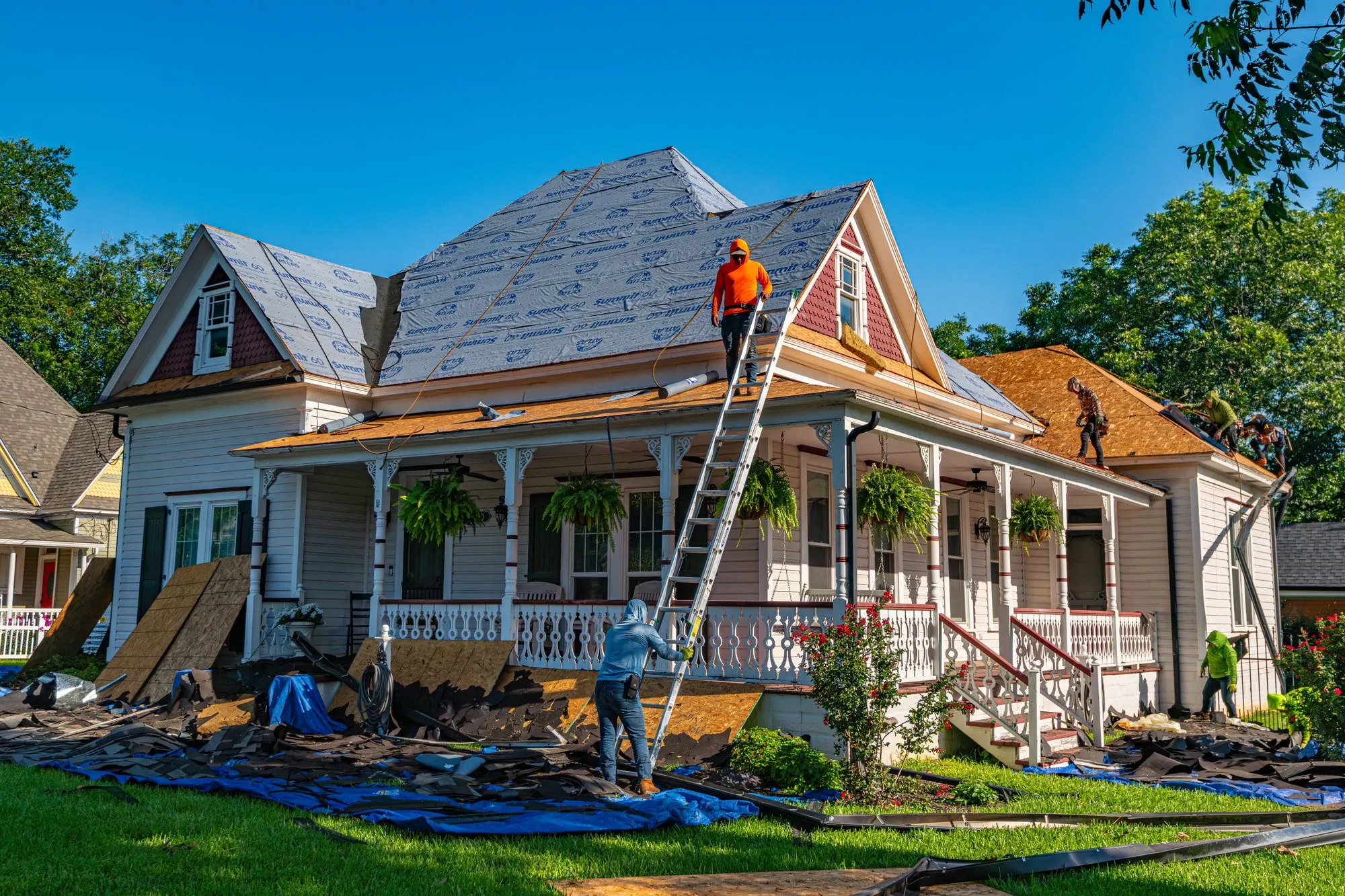 A ladder set against a porch roof as a crew re-covers a storm-damaged house